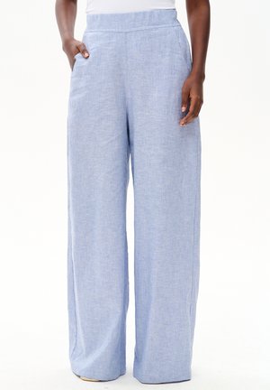 Kobieta nosząca jasnoniebieskie, luźne spodnie lniane z wysokim stanem i bocznymi kieszeniami, stojąca na białym tle.