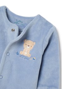Pelele de bebé de felpa azul claro con un oso bordado y acentos florales, con cierres a presión en la parte delantera. Textura suave.