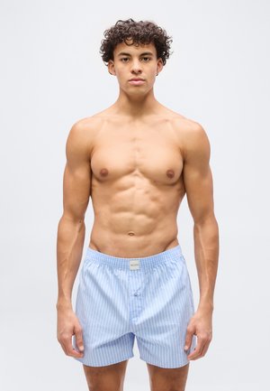Ung mann med krøllete hår står skjorteløs iført lyseblåstripede boxershorts, vendt mot fronten mot en ensfarget bakgrunn.