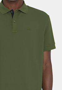 Polo verde oliva realizzato in tessuto morbido, con patta a due bottoni e un logo sottile dello stesso colore sul petto.