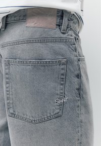 Gros plan du jean en denim gris avec une petite poche arrière brodée du logo « Supreme » et une étiquette en cuir marron sur la ceinture.