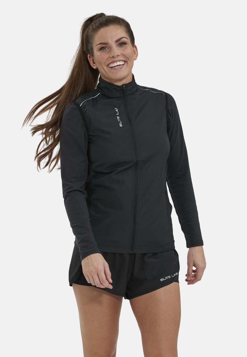 Ung kvinde smilende og stående, iført sort atletisk vest med lynlås og matchende shorts med "Elite Lab" logoer.