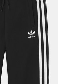 Černé tepláky z bavlny s bílými detaily v podobě tří pruhů po stranách a malým bílým vyšitým logem Adidas na přední straně.