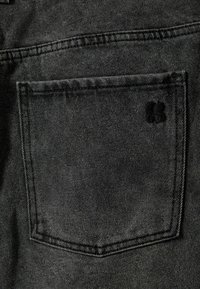 Črn jeans material v bližnjem načinu, ki prikazuje zadnji žep z izvezenim robom in vtisnjenim logotipom v kotu. Površinska tekstura je subtilno vidna.