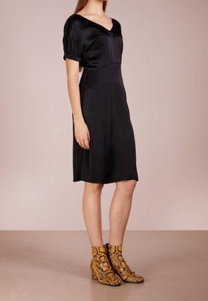 Kobieta w czarnej, satynowej sukience do kolan z bufkami i żółtymi botkami z wężowej skóry stojąca na posadzce w cętki.