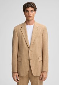 Beige gestreifter Blazer aus strukturiertem Stoff, mit Reverskragen, zwei Knöpfen und seitlichen Taschen. Über einem weißen T-Shirt getragen.