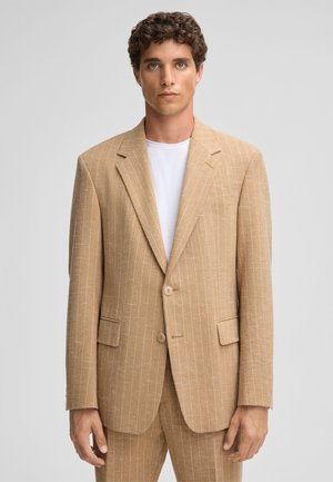 Tans gestreepte blazer van textuurstof, met notch-lapels, twee knopen en zijzakken. Draagt over een wit T-shirt.