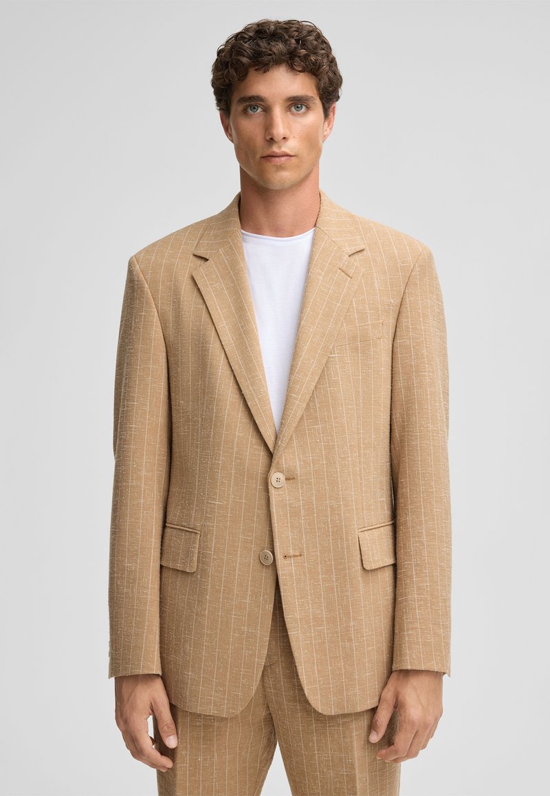 Beige gestreifter Blazer aus strukturiertem Stoff, mit Reverskragen, zwei Knöpfen und seitlichen Taschen. Über einem weißen T-Shirt getragen.