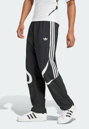 Mężczyzna noszący czarne spodnie dresowe Adidas z białymi paskami i logo, w połączeniu z białymi butami sportowymi Adidas z czarnymi paskami.
