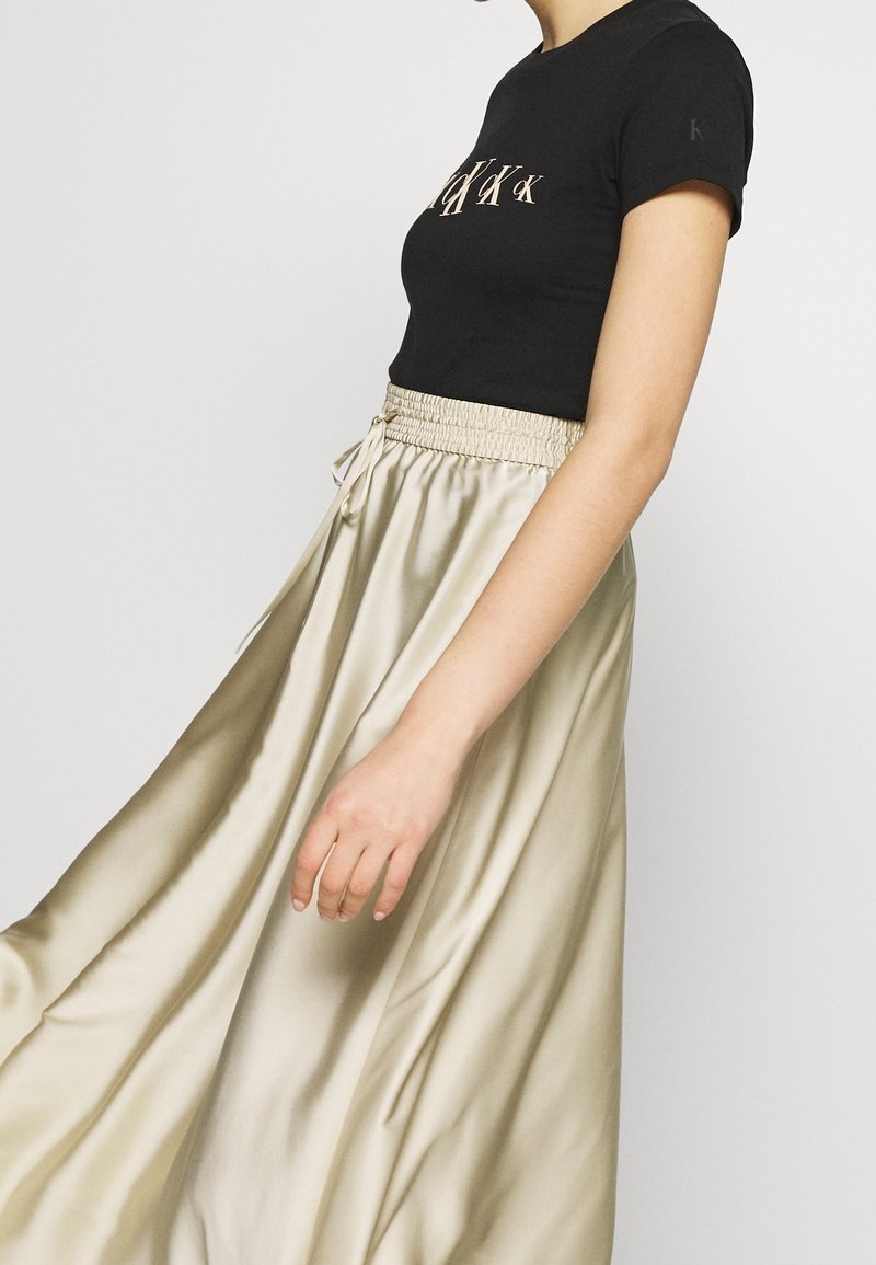 Kobieta ubrana w czarny dopasowany T-shirt z jasnym logo i zwiewną beżową satynową spódnicę, stojąca na tle jednolitego jasnego tła.