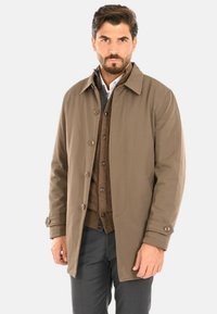 Giacca impermeabile marrone con chiusura a bottoni, colletto classico e tasche laterali, indossata sopra un maglione e una camicia marroni.