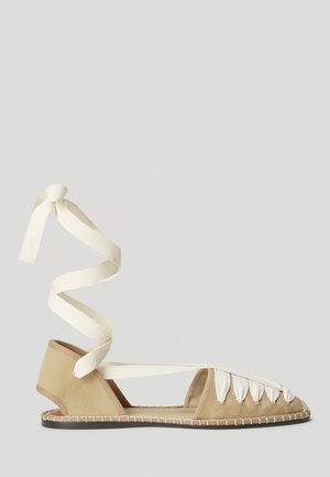 Zapato plano de lona color beige con un cordón blanco en espiral, lados abiertos y cordones decorativos en la parte frontal. Textura suave y detalles de costura contrastante.
