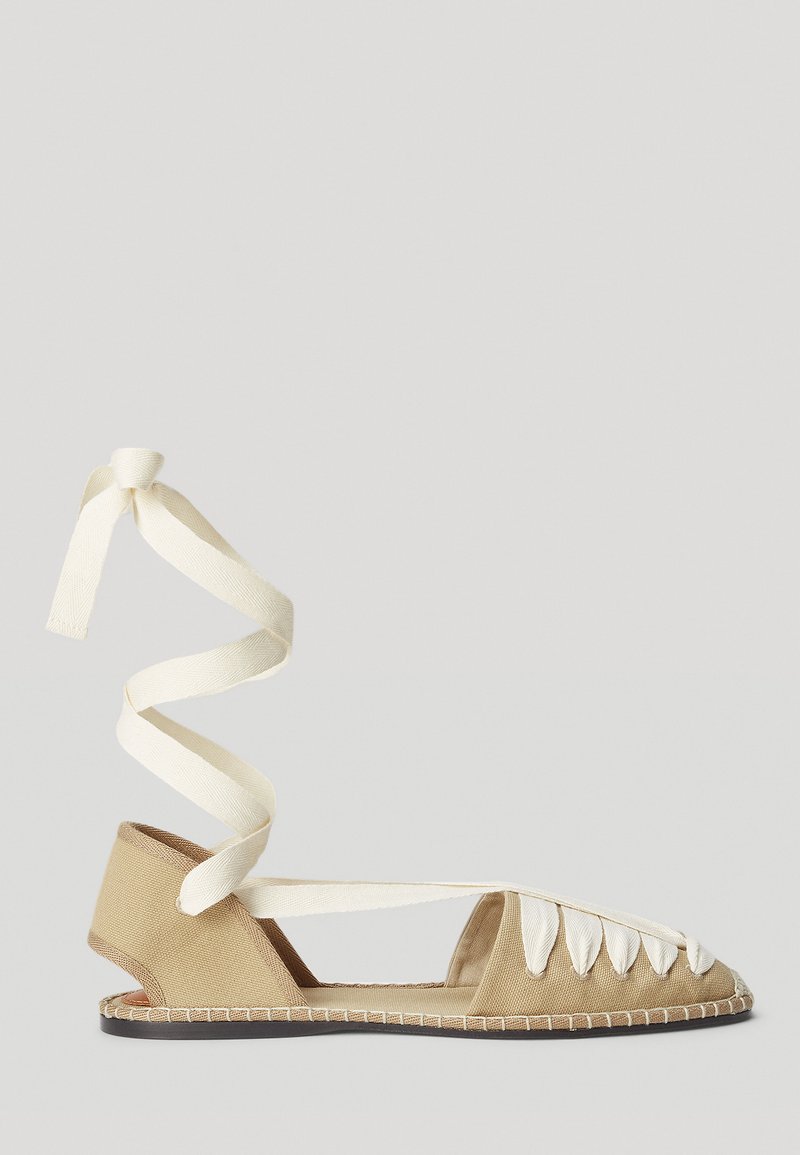 Tan canvas flat shoe with a spiral white tie, open sides, and decorative lacing across the front. Soft texture and contrast stitching detail.