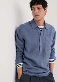 Vaaleansininen pitkähihaine paita pellava-sekoitetta, jossa on nappikaulus, rintatasku ja vaalean- ja mustanruskeat raidalliset hihansuut.