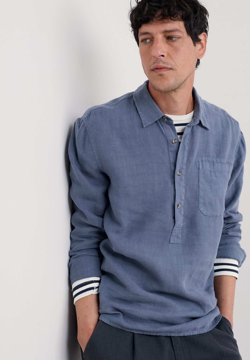 Vaaleansininen pitkähihaine paita pellava-sekoitetta, jossa on nappikaulus, rintatasku ja vaalean- ja mustanruskeat raidalliset hihansuut.