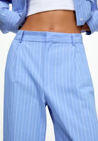 Kobieta ubrana w jasnoniebieskie spodnie w paski z wysokim stanem oraz pasującą marynarkę, pod którą ma białą krótki top, widoczny tors.