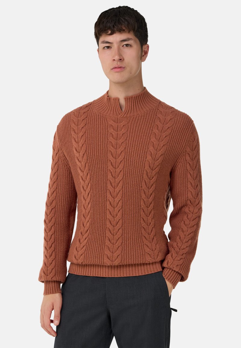 Pull en maille torsadée marron avec col montant côtelé, motif texturé, poignets et ourlet côtelés. Associé à un pantalon foncé.