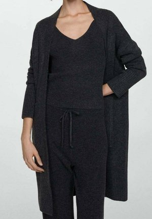 Frau trägt eine dunkelgraue Strickjacke, ein passendes ärmelloses V-Ausschnitt-Oberteil und eine Hose mit Kordelzug, posiert mit einer Hand in der Tasche vor weißem Hintergrund.