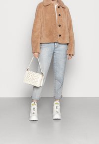 Giacca in finta pelliccia marrone con ampio colletto e bottoni neri, abbinata a jeans in denim chiaro e sneakers bianche con lacci. Borsa intrecciata bianca.