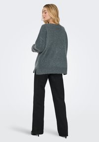 Prevelik dolg pulover iz sive pletenine z rebrastim robom, stranskimi razporki in ramenskimi šivi v padajoči liniji, kombiniran s črnimi širokimi hlačami in visokimi petami.