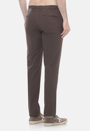 Uomo che indossa pantaloni marrone scuro e sneakers marroni, in piedi a piedi nudi contro uno sfondo chiaro e uniforme, visto da dietro.
