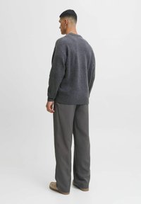 Grauer Strickpullover mit geripptem Saum, getragen über lockeren, grauen Hosen. Das Model steht mit dem Rücken zur Kamera und zeigt die entspannte Passform und Textur des Pullovers.