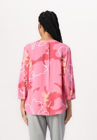 Rosa Bluse mit großem Blumenmuster in Rottönen, Creme und Weiß, mit Dreiviertelärmeln und einem leicht abgerundeten Saum.