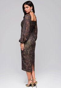 Schwarzes, blumenbedrucktes Kleid mit langen, gepufften Ärmeln, einem quadratischen Ausschnitt und einem seitlichen Schlitz. Ein Paar olivgrüne Absatzsandalen vervollständigt den Look.
