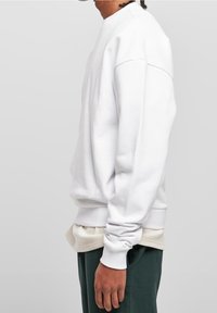 Vit crewneck-tröja med ribbade muddar och fåll, mjuk textur, bärs över ett beige lager-på-lager-plagg, mot en neutral bakgrund.