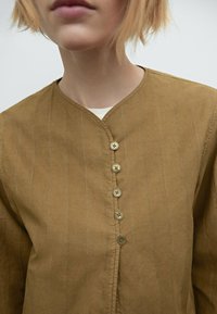 Bruine top met knopen en een V-hals, voorzien van verticale witte strepen en vijf lichtgekleurde knopen aan de voorkant. Gladde stoftextuur.