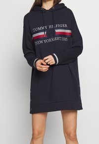 Marinblå hoodie-klänning med långa ärmar, snörning i huvan, logotyp framtill och knappdetaljer på sidan. Mjuk tyg med ribbade manschetter och fåll.