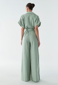 Zeleni širok kombinezon z kratkimi zavihanimi rokavi, z pasom in lahkotnim materialom. Ima gladko teksturo in podrobnost na hrbtnem delu.