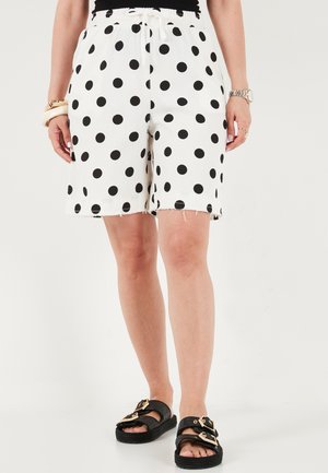 Kvinde iført hvide shorts med sorte prikker, sort top uden bælte, sorte sandaler med guldlåse, ur og armbånd på hvid baggrund.