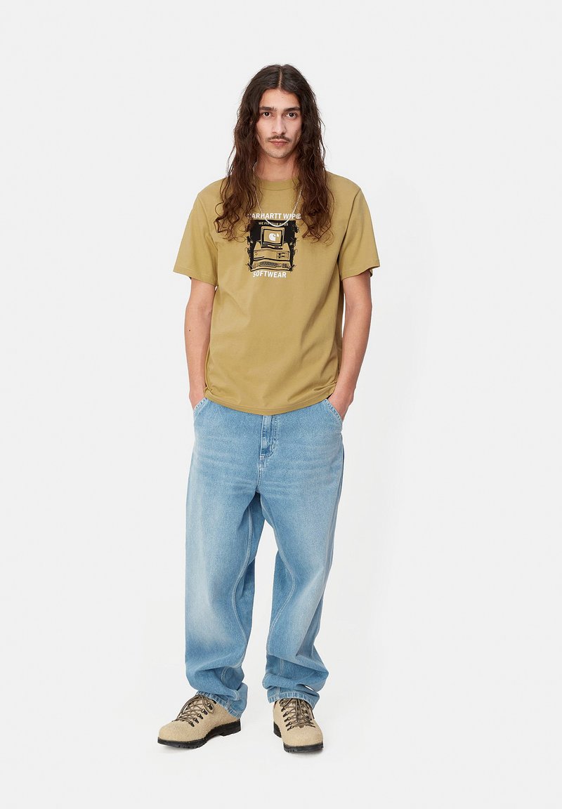 Camiseta caqui con estampado gráfico, corte holgado. Pantalones vaqueros holgados azul claro, combinados con zapatos beige con cordones. Cabello largo castaño, manos en los bolsillos.
