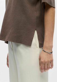 Bras droit avec bracelet argenté, portant un haut marron à manches courtes et un pantalon crème avec poche latérale, debout devant un fond blanc.