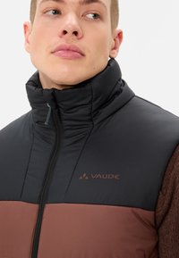 Herrenweste in Schwarz und Braun, mit hohem Kragen, Reißverschluss und Logodetail. Strukturierter Stoff mit kontrastierenden Farbbereichen.