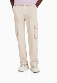 Beige cargo kalhoty se dvěma bočními kapsami a dvěma předními cargo kapsami. Vyrobené z odolného materiálu, volný střih a rovné nohavice.