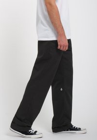 Pantalones holgados negros de mezcla de algodón con un detalle de logo en la pierna, combinados con zapatillas negras y blancas y una camiseta blanca.