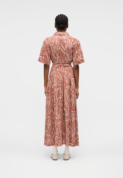 Persona con cabello trenzado, vestida con un vestido floral color óxido con cinturón y mangas cortas, de pie de espaldas frente a un fondo liso.
