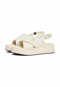 Platform sandalen van crème synthetisch materiaal. Kruisende banden, verstelbare enkelband, vlakke zwarte zool, met reliëflogo-detail aan de zijkant.