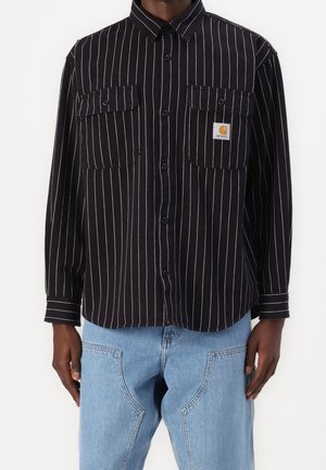 Zwart overhemd met lange mouwen en knopen, witte verticale strepen, twee borstzakken met knopen, inclusief een Carhartt-logopatch, gedragen met lichtblauwe spijkerbroek.