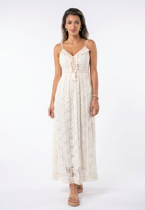 Woman wearing a sleeveless cream lace maxi dress with floral patterns and beige open-toe sandals, standing against a white background.