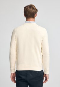 Sudadera de color crema claro con mangas largas, puños acanalados y una textura suave, combinada con pantalones oscuros. La vista trasera muestra un diseño limpio.