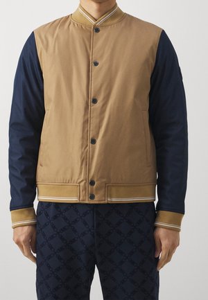 Persona che indossa una giacca bicolore con parte anteriore color cuoio e maniche blu, abbottonata, e pantaloni blu scuro con un motivo geometrico sottile.