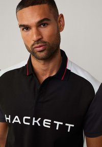 Camisa polo negra con el logotipo blanco "HACKETT", mangas en blanco y gris oscuro en contraste, y un collar abotonado con acentos rojos.