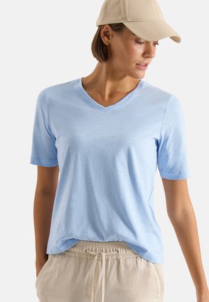 Kvinde iført en lyseblå T-shirt med V-hals, beige bukser med snøre og en beige baseballkasket, der kigger ned mod sin højre side.