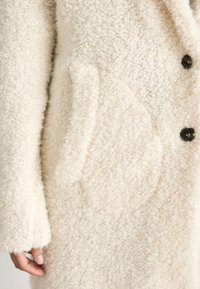White fleece coat with a soft, textured finish, featuring a large, subtly patterned pocket and black button accents.