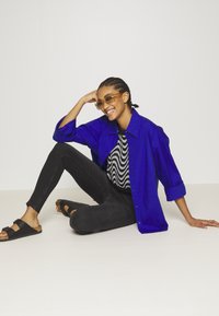 Camisa oversized azul, suéter preto com padrão ondulado, jeans pretos, sandálias pretas. Modelo sentado com um fundo claro e a sorrir.