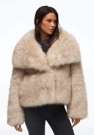 Femme portant une veste en fausse fourrure beige, un haut gris, un pantalon noir avec des fentes à l'avant, et des chaussures noires à talons, debout devant un fond blanc.
