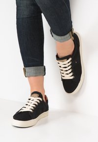 Sneakers a rete nera con lacci crema e suola in gomma bianca. Presentano dettagli dorati e una superficie testurizzata. I jeans sono arrotolati ai polsi.